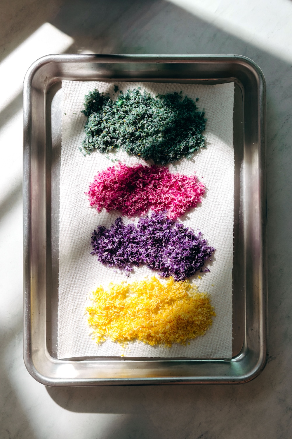 Vegan Sprinkles | DIY Sugar Free Recipe Four piles of naturally colored coconut sprinkles on parchment paper in a baking pan — green, pink, purple, and yellow — before drying