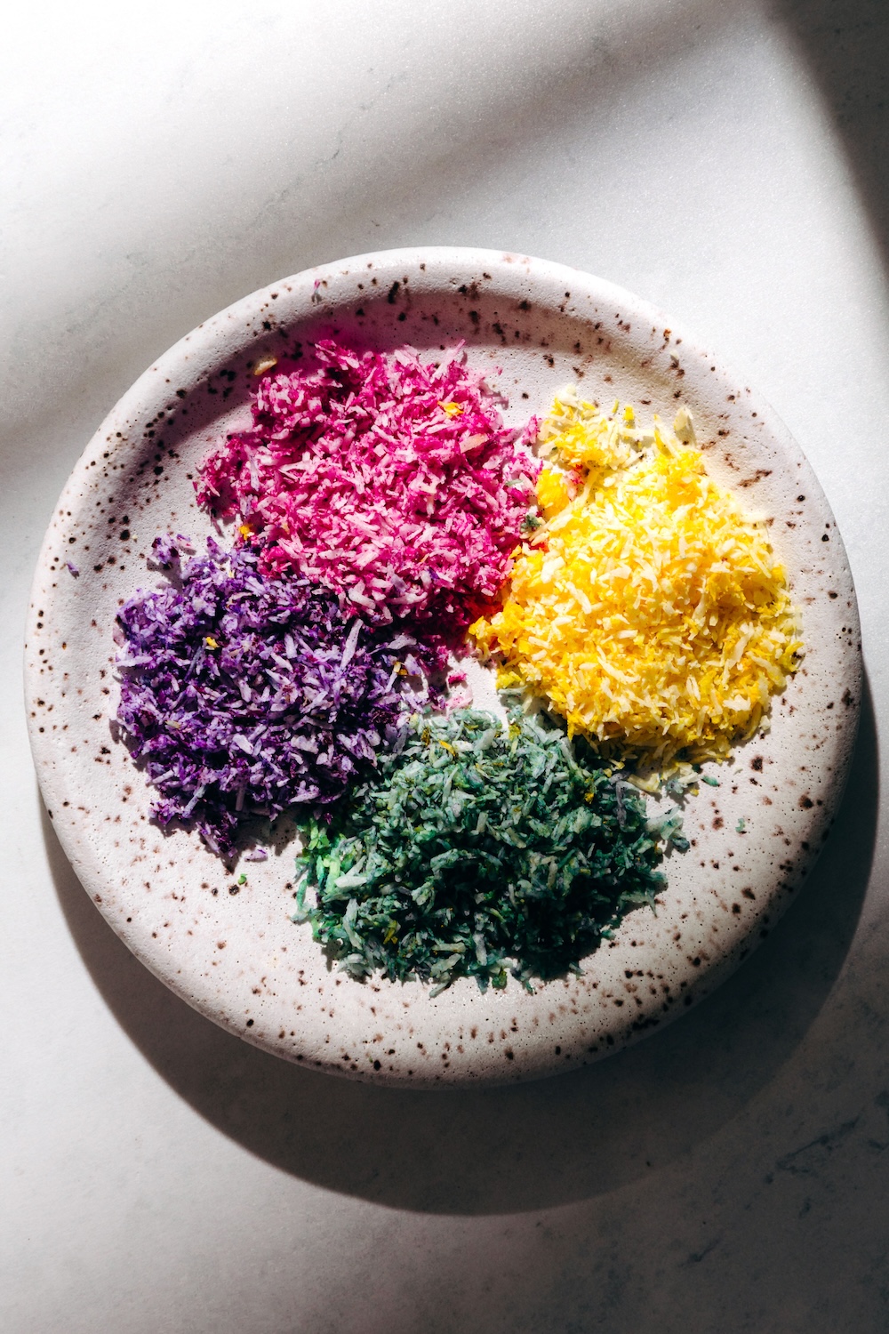 Vegan Sprinkles | DIY Sugar Free Recipe Four separate piles of homemade dye-free coconut sprinkles in a speckled ceramic bowl — pink, yellow, purple, and green