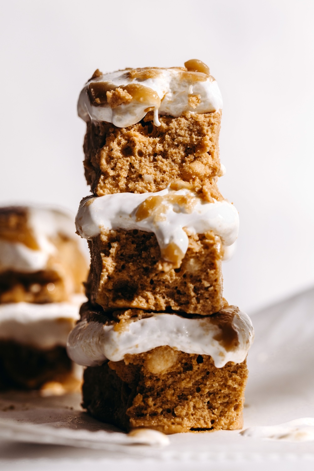 Three vegan fluffernutter protein bars stacked tall on parchment paper, with marshmallow fluff and golden SunButter caramel drizzling down the sides against a light background