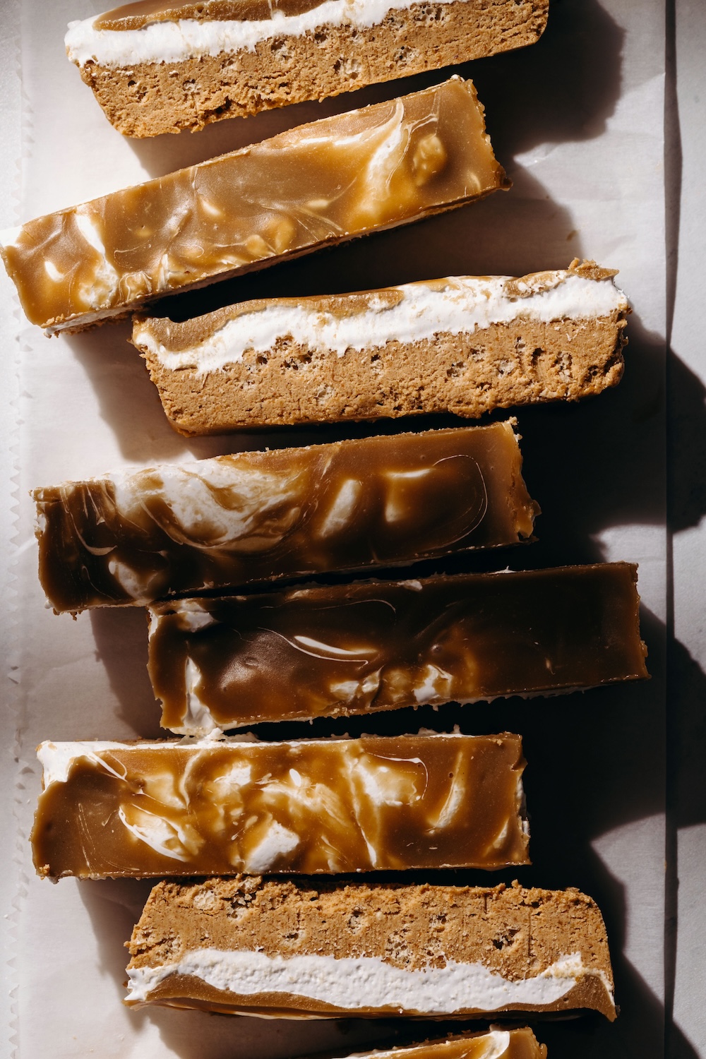 Overhead view of sliced vegan fluffernutter protein bars arranged on parchment paper, each showing a golden SunButter base, white marshmallow layer, and glossy swirled chocolate caramel topping
