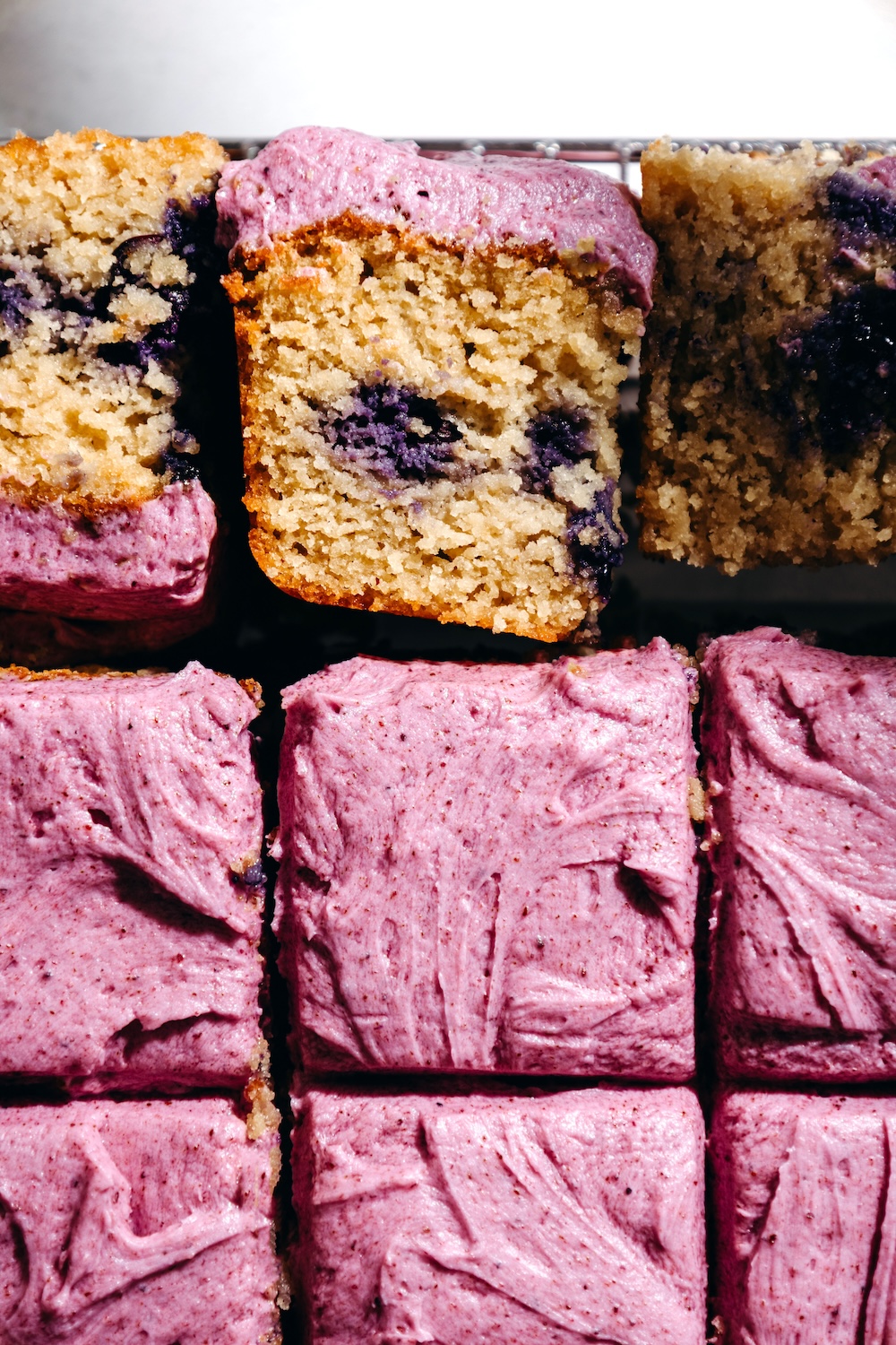 Gluten free blueberry snack cake squares topped with pink blueberry frosting, one slice tipped on its side showing the moist crumb studded with blueberries.