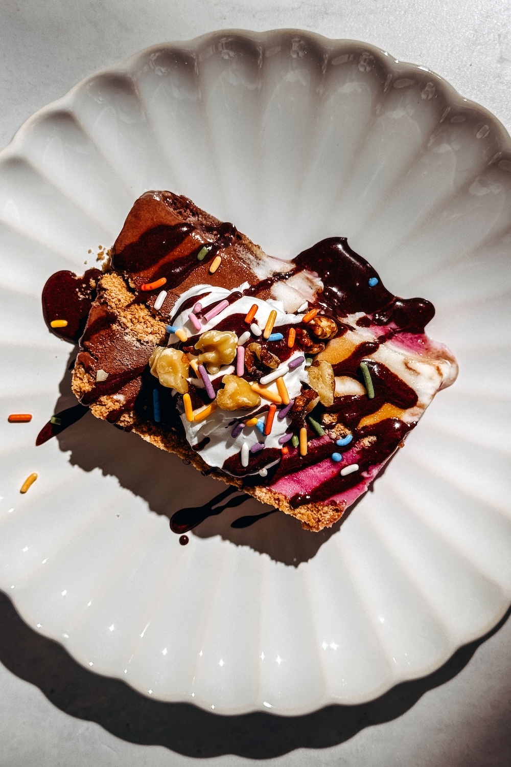 Overhead view of a no-churn loaf pan ice cream cake slice topped with whipped cream, sprinkles, walnuts, and chocolate sauce on a ceramic plate