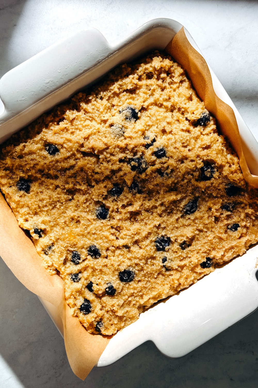 Unbaked gluten-free blueberry snack cake batter studded with blueberries in a parchment-lined square baking dish.