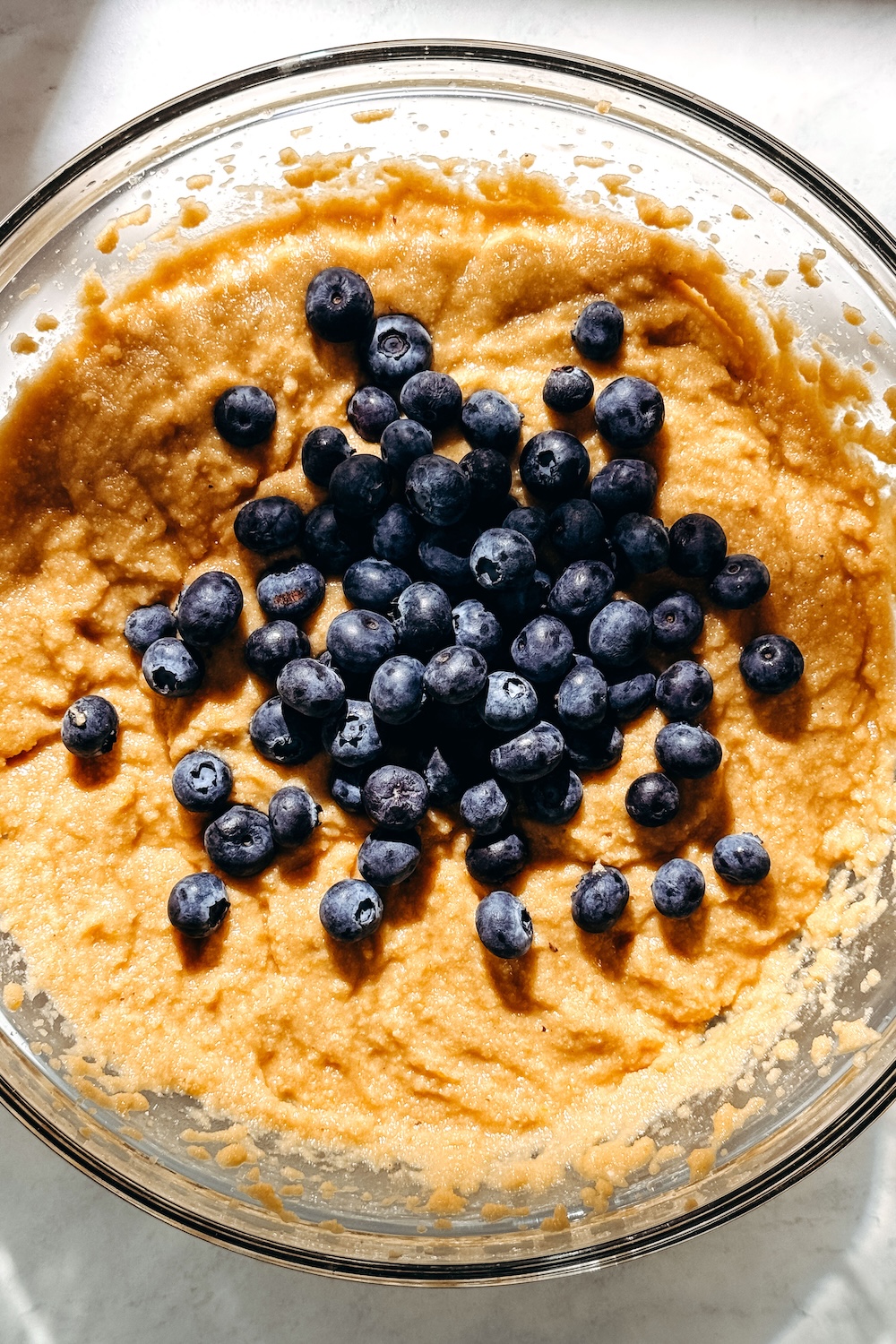 Fresh blueberries piled on top of cake batter in a glass mixing bowl before being folded in.