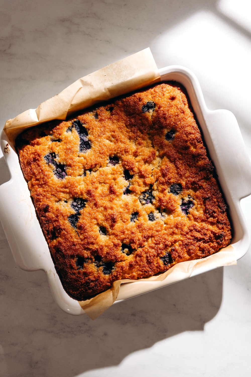 Baked gluten-free blueberry snack cake in a square dish lined with parchment paper, golden brown top studded with blueberries.