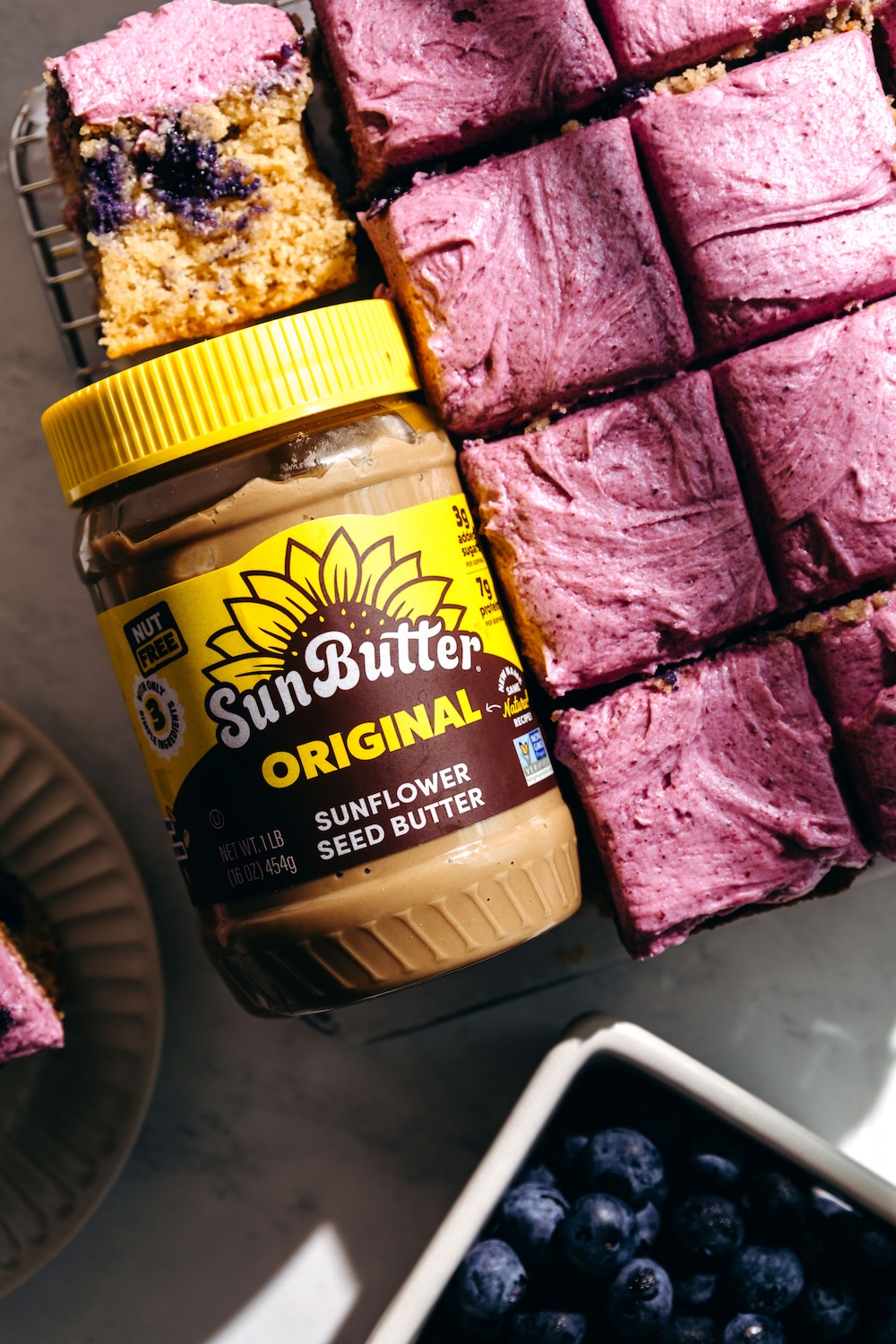 A jar of Original SunButter sunflower seed butter next to sliced gluten-free blueberry snack cake with fresh blueberry frosting and a bowl of blueberries.