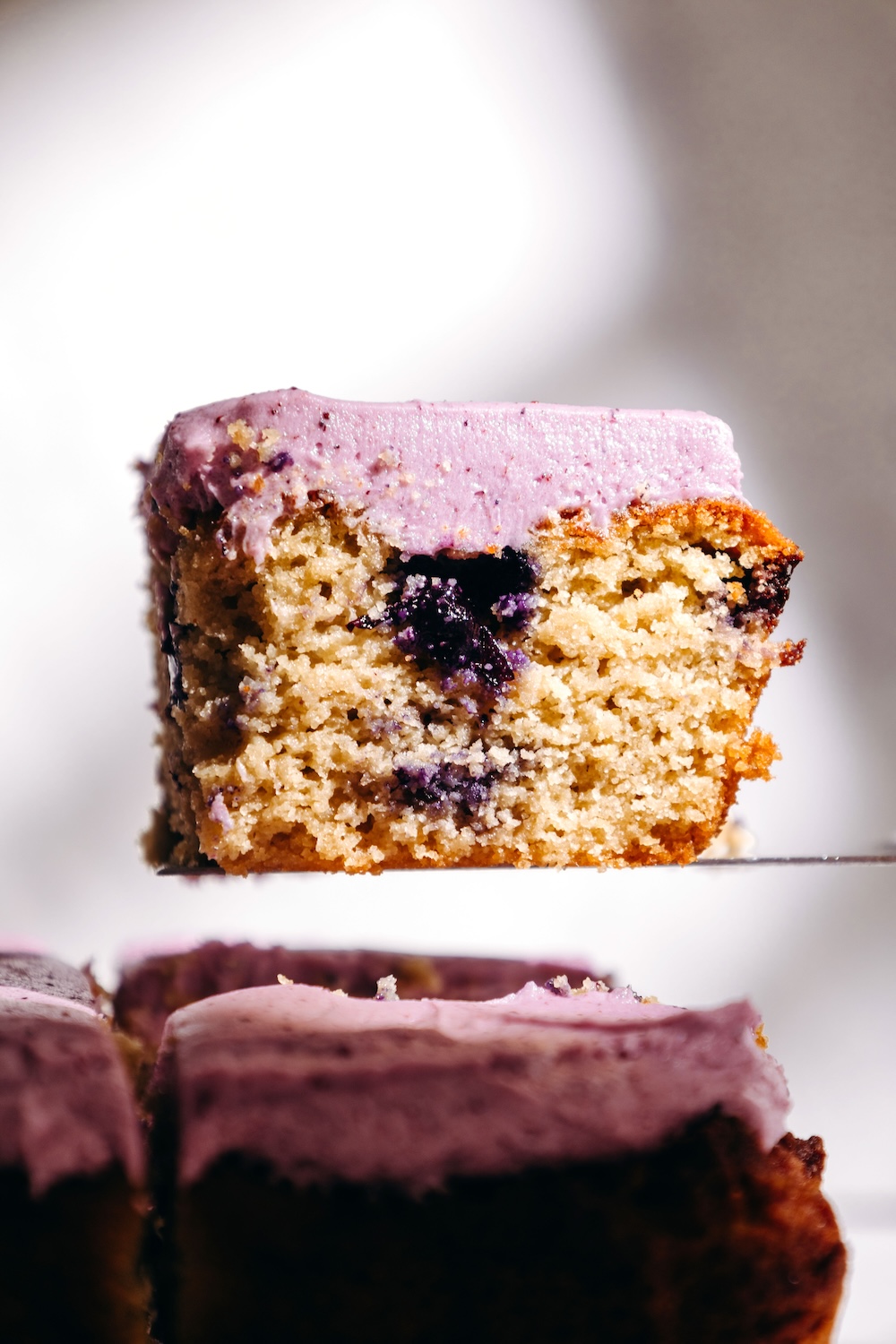 Close-up of a frosted gluten-free blueberry snack cake slice balanced on a knife, showing the crumb and a pocket of baked blueberries.