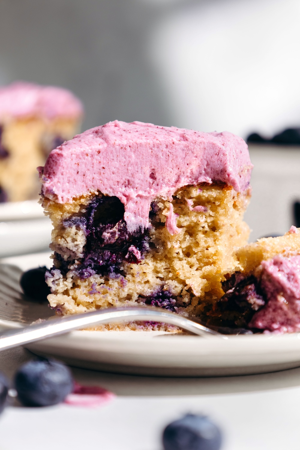 Close-up of a slice of gluten-free blueberry snack cake with fresh blueberry frosting, a fork resting beside it on the plate.