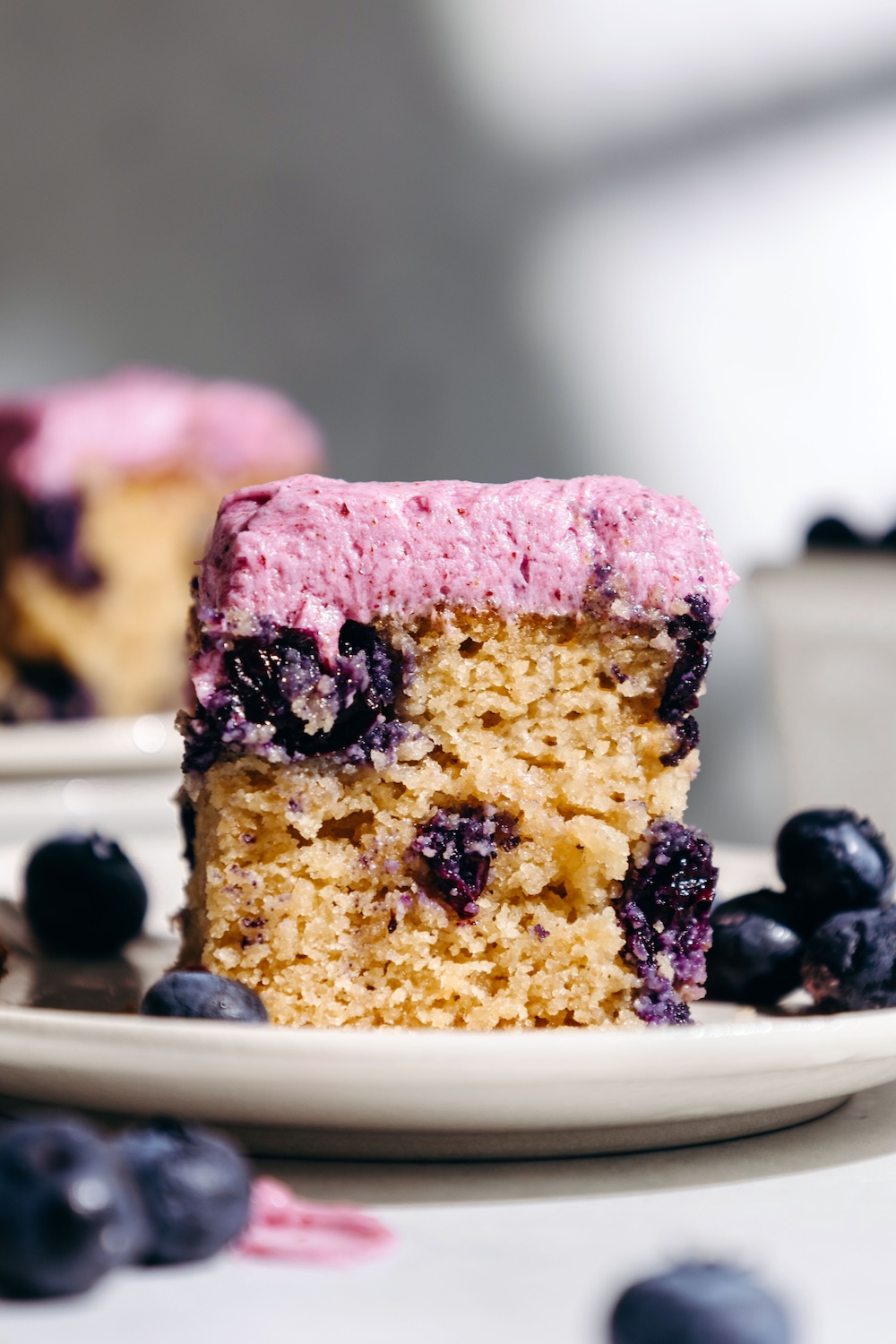 Slice of gluten-free blueberry snack cake with fresh blueberry frosting on a plate, surrounded by fresh blueberries with a second frosted cake blurred in the background.