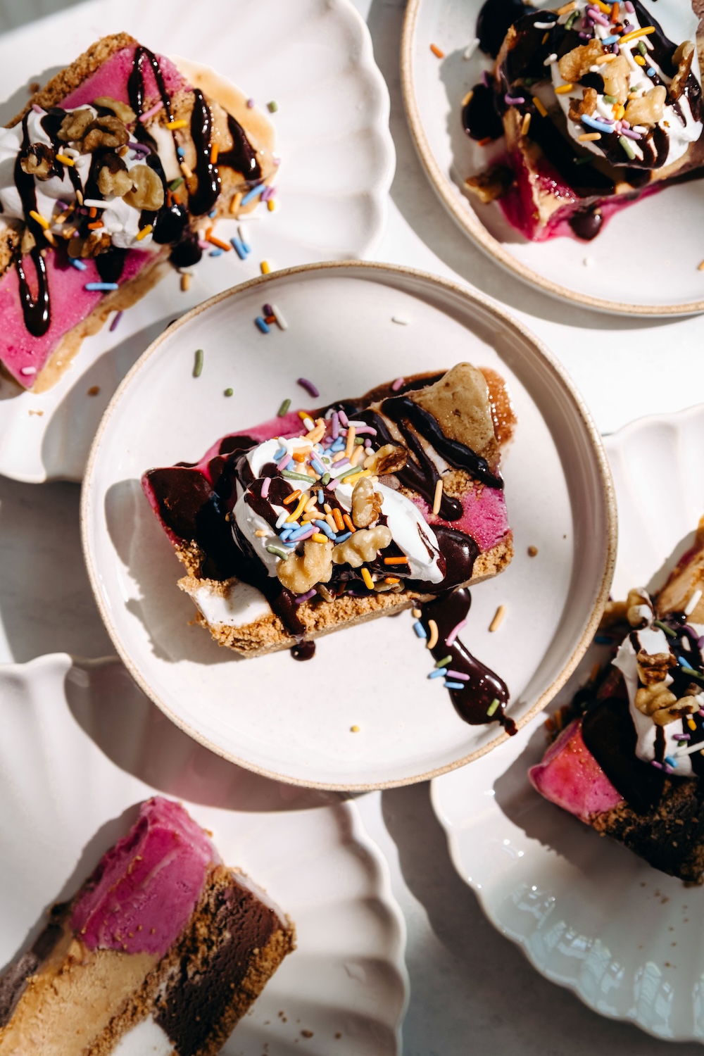 Slices of loaf pan ice cream cake with sugar cone crust topped with whipped cream, sprinkles, walnuts, and chocolate sauce on white plates