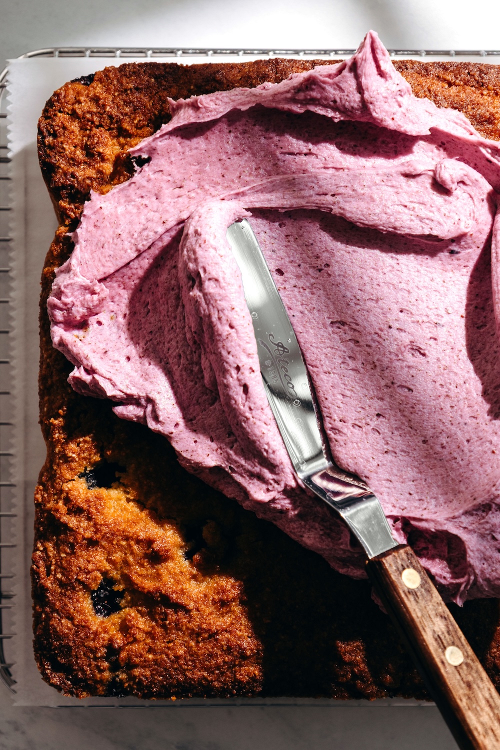 Fresh blueberry frosting being spread over a baked gluten-free blueberry snack cake with an offset spatula.