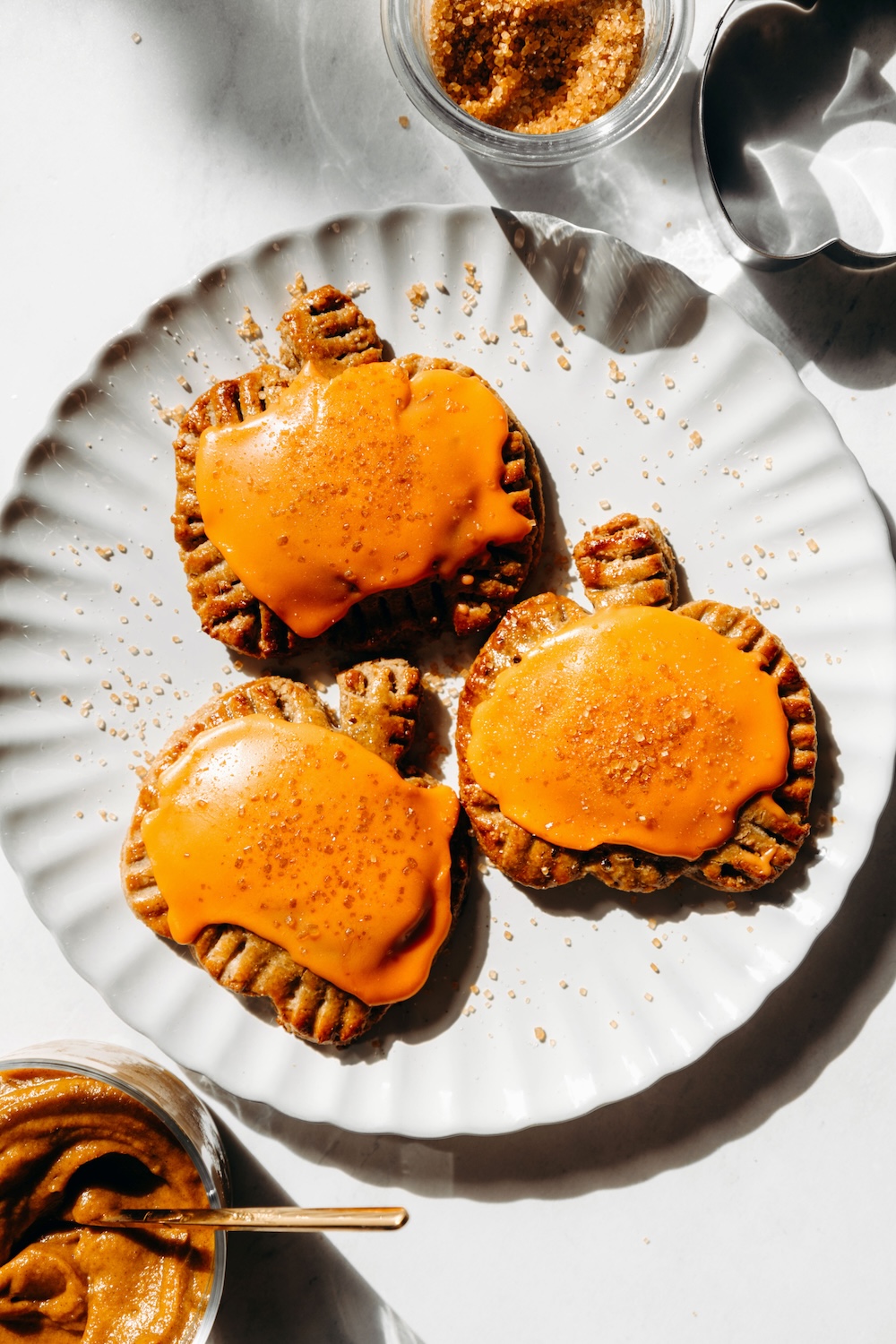 Overhead view of three pumpkin-shaped pop tarts with bright orange glaze on a white scalloped plate, with a bowl of pumpkin filling and a pumpkin cookie cutter nearby.