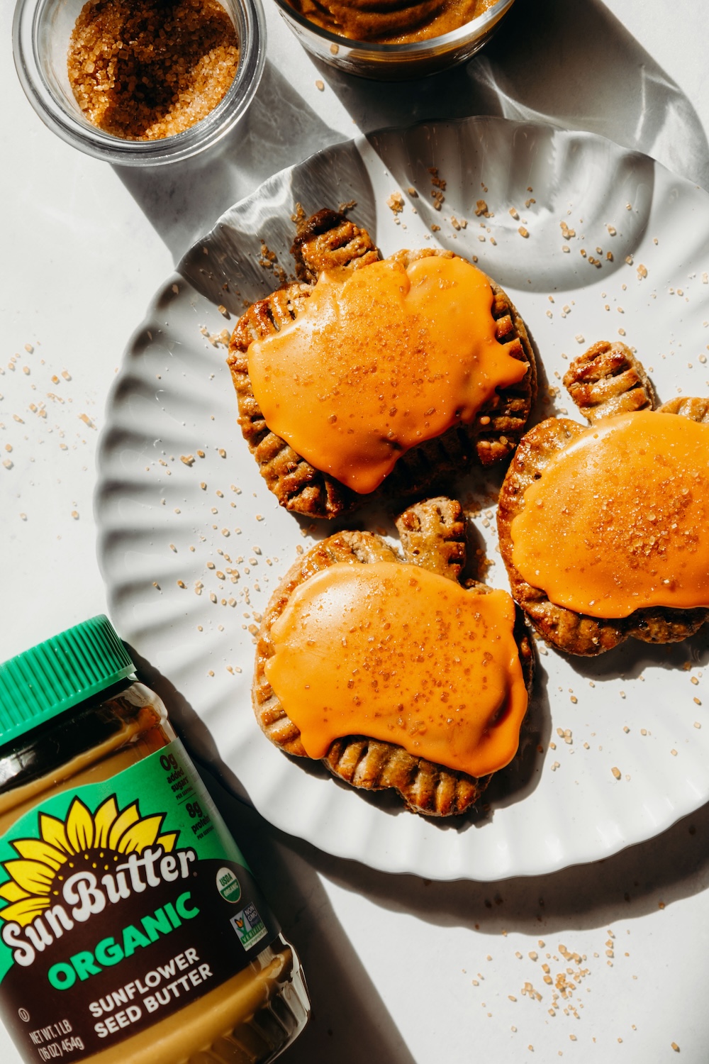Three gluten-free pumpkin pie pop tarts with orange glaze and sanding sugar on a white scalloped plate, next to a jar of SunButter Organic Sunflower Seed Butter.